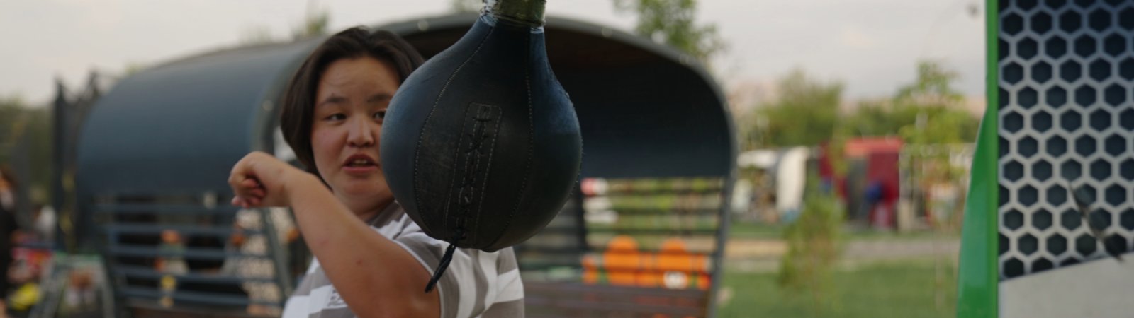 A girl swings back to hit a black boxing ball. She is wearing a striped shirt, is slightly overweight and looks rather Mediterranean with dark hair. She is standing outdoors, behind her is a small square with a covered seating area. The background is blurred and looks more like a campsite. The girl is a teenager and is looking intently at the ball.