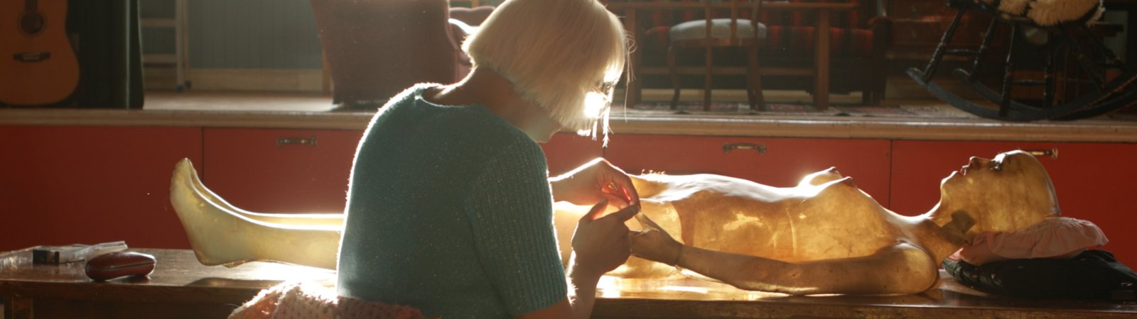 A woman wearing a turquoise shirt and black shorts is seen from the side, from behind. She is sitting on a chair in front of a long wooden table on which a life-size female figure is lying. Old-fashioned furniture and a window can be seen in the background. The scene is lit by daylight, which illuminates the slightly transparent figure. There is a quiet, concentrated atmosphere.