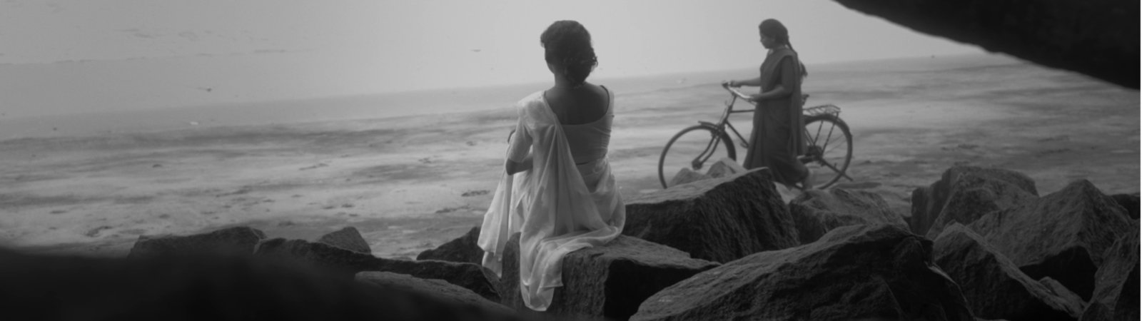 A black-and-white image with light, soft contrasts showing a woman in a sari sitting between rocks. She is sitting with her back to the viewer and looking out to sea. A long wave rolls in. To her right stands another woman in a sari, holding a bicycle and also looking out to sea.
