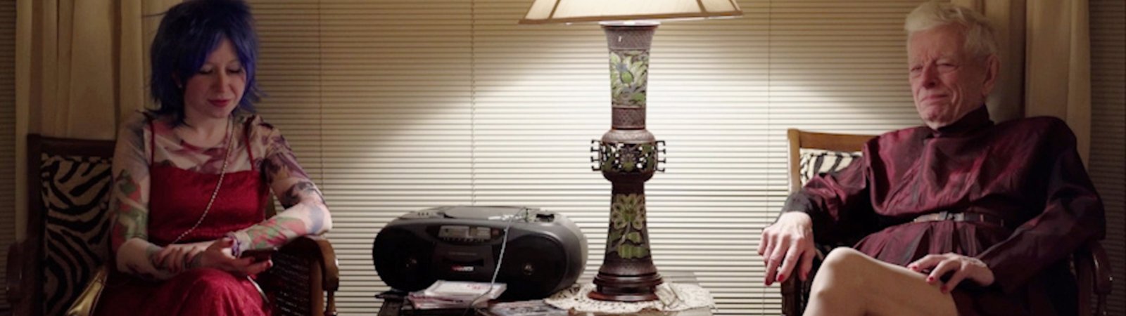 The scene shows two people sitting in front of a lowered blind. A cone of light emanates from a large lampshade lamp standing on a side table in the centre of the picture. Next to the lamp is a portable CD player. To the left of the table sits a younger woman with blue hair, wearing a striking red dress and high red shoes. She has her legs crossed and her gaze lowered, as if she is thinking or waiting. To the right of the table, sitting slightly diagonally, is a white-haired gentleman wearing a dark red top and with bare legs. He also has his legs crossed and is looking straight ahead. The whole situation seems to be one of waiting.